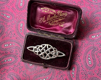 May include: A silver-colored Celtic knot brooch displayed in an open, burgundy velvet-lined jewelry box. The box is embossed with gold lettering, including the name "A. Marks & Sons". The background is a pink paisley pattern.