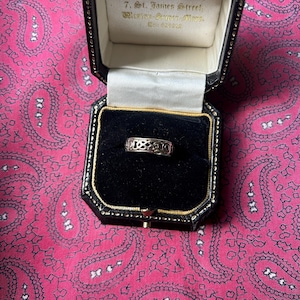 May include: A silver ring with an openwork design, displayed in a black jewelry box with a gold interior. The box is open, revealing the ring's intricate details. The background is a pink paisley fabric.