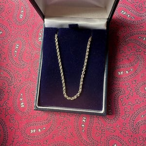 May include: A gold-colored chain necklace displayed in a black box with a blue velvet interior. The box is open, revealing the necklace against the dark blue background. The box sits on a red paisley patterned surface.