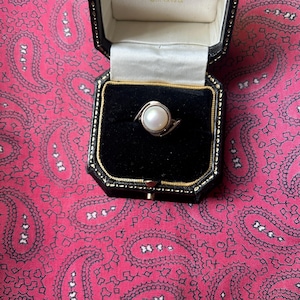 Puede incluir: Un anillo de plata con una perla blanca grande y redonda en un diseño en espiral. El anillo se muestra en una caja de joyería negra con interior de satén blanco, sobre un fondo rojo con estampado de cachemira.