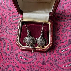 May include: A pair of silver-colored turtle earrings displayed in a red velvet-lined jewelry box. The earrings have a detailed, textured design. The box is open, revealing the earrings against the plush interior. The background is a pink paisley pattern.