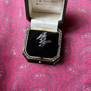 Puede incluir: Un anillo de plata con un diseño único, con múltiples piedras preciosas ovales de color azul oscuro. El anillo se exhibe en una caja de joyería negra con ribetes dorados, sobre un fondo rosa con estampado de cachemira.