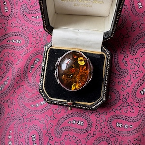 Puede incluir: Un anillo ovalado de ámbar con engaste de plata, exhibido en una caja de joyería negra con interior blanco. La piedra de ámbar tiene un tono marrón dorado cálido con inclusiones visibles. El anillo se encuentra sobre un fondo rosa con estampado de cachemira.