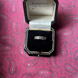 May include: A silver ring with a repeating openwork design, displayed in a black jewelry box with gold trim. The box is open, revealing the ring against a black velvet interior. The background is a pink paisley pattern.