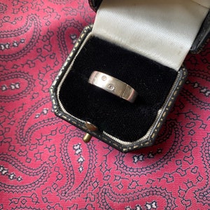 May include: A silver band with two small diamonds set into the band. The ring is in a black velvet ring box.