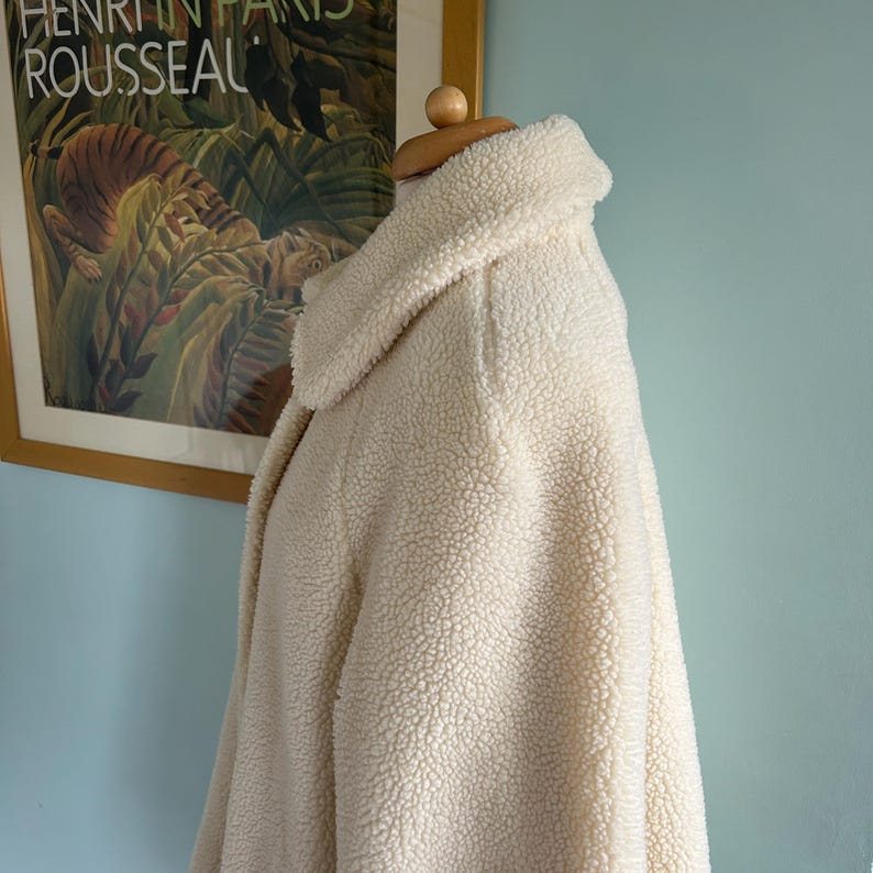 May include: A cream-colored faux fur jacket with a high collar and long sleeves. The jacket is made of a soft, fluffy material and has a relaxed fit.