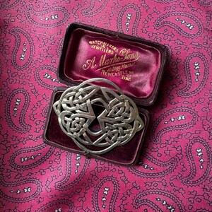 May include: A silver-toned Celtic knot brooch displayed in a vintage maroon jewelry box with gold lettering. The brooch features an intricate interwoven design. The box is on a pink paisley patterned fabric.