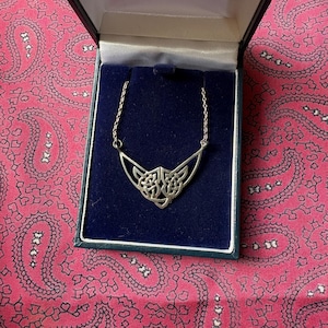 May include: A silver-toned Celtic knot pendant necklace displayed in a blue velvet-lined box. The necklace features a detailed, symmetrical knot design at the center, with a V-shaped frame. The box is set against a pink paisley patterned background.