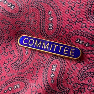 Op de afbeelding: Een blauwe en gouden emaille pin met het woord "COMMITTEE" in witte letters.
