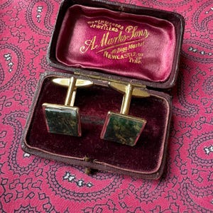 Puede incluir: Un par de gemelos dorados con incrustaciones cuadradas de piedra veteada verde y marrón, presentados en un estuche marrón con bisagras forrado de terciopelo rojo oscuro. La tapa interior muestra el texto dorado A. Marks & Sons.