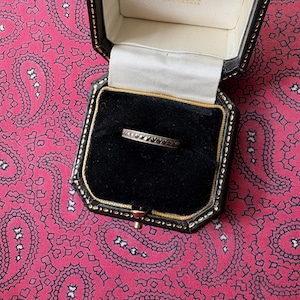 Puede incluir: Un anillo de plata con un patrón repetido de pequeñas piedras oscuras se muestra dentro de un joyero negro con un borde dorado. La caja está abierta, revelando el anillo contra un interior de terciopelo negro. El fondo es un estampado de cachemira rosa.