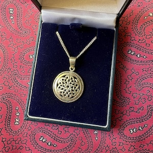 May include: A silver-toned circular pendant featuring an intricate openwork Celtic knot design with a twisted rope border, suspended from a matching silver-toned chain. The necklace is presented inside a dark blue velvet-lined jewelry box.