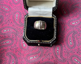 Könnte beinhalten: Ein silberner Ring mit rechteckigem Design, präsentiert in einer schwarzen Schmuckschatulle mit goldenem Rand. Der Ring hat eine vertikale Reihe kleiner, klarer Steine. Die Schatulle ist geöffnet und zeigt einen schwarzen Samtinnenraum.