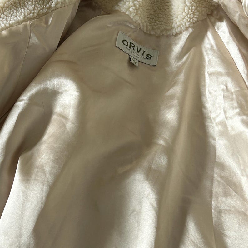 May include: A cream-colored jacket with a soft, fuzzy lining. The jacket has a label that reads "ORVIS" and "Made in China".