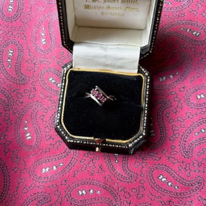 May include: A silver ring with three pink gemstones set in a diagonal design, displayed in a black jewelry box with gold trim. The box is open, revealing the ring against a black velvet interior. The background is a pink paisley pattern.