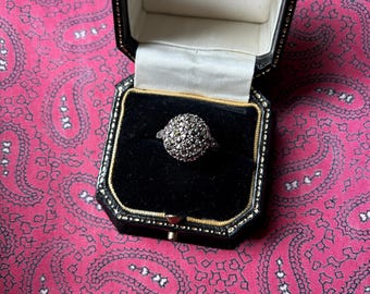 Puede incluir: Un anillo de plata con un grupo de pequeñas piedras redondas engastadas en forma de cúpula, expuesto en una caja de joyería negra con ribete dorado. La caja está abierta, revelando el anillo contra un interior de terciopelo negro. El fondo es un estampado de cachemira rosa.