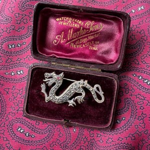 May include: A silver dragon brooch with a red gemstone eye sits in an open, burgundy velvet-lined jewellery box. The box is embossed with gold lettering, reading "A. Marks & Sons, Watchmakers & Jewellers, 40 Bigg Market, Newcastle on Tyne."