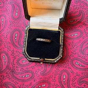 Puede incluir: Un delicado anillo con pequeñas piedras brillantes engastadas en una banda dorada, presentado en una caja de joyería negra con ribete dorado. La caja está abierta, revelando el anillo contra un interior de terciopelo negro. El fondo es un estampado de cachemira rosa.