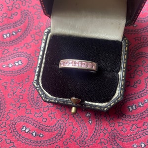 Puede incluir: Un anillo de plata con una fila de piedras preciosas cuadradas de color rosa engastadas en una banda.