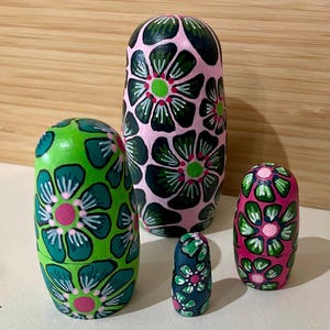 May include: A set of five wooden nesting dolls, each painted with a floral design. The dolls are in shades of green, pink, and blue, with white accents. The largest doll is approximately 6 inches tall.