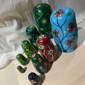 May include: A collection of hand-painted Russian nesting dolls, or Matryoshka dolls, in various sizes. The dolls feature floral designs in shades of green, blue, red, and yellow. The dolls are arranged on a light wooden surface.