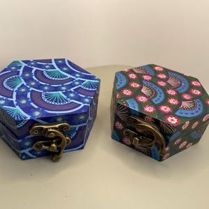 Peut inclure: Deux boîtes hexagonales en bois avec des couvercles décoratifs. La boîte de gauche est bleue avec un motif en éventail dans les tons de bleu et de violet. La boîte de droite est verte avec un motif floral rose et bleu. Les deux ont des fermoirs en laiton.