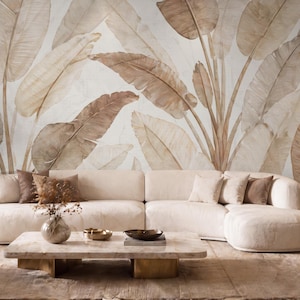 May include: A large, cream-colored sectional sofa sits in front of a wall mural featuring oversized, muted brown banana leaves. The room includes a low, rectangular stone coffee table, a matching armchair, and a side table with a lamp.