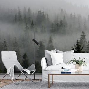 May include: A white couch with gray pillows sits in front of a wall mural depicting a foggy forest. A gray chair with a white throw blanket sits to the left of the couch. A glass coffee table with books and a potted plant sits in front of the couch.