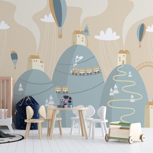 May include: A nursery room with a light blue and beige wallpaper featuring a whimsical landscape with mountains, houses, and hot air balloons. The room includes a cot, a toy wagon, and a table with chairs.