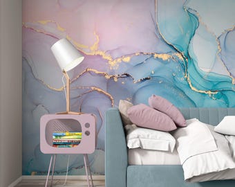 May include: A wall mural featuring an abstract fluid art design in shades of pastel purple, blue, and teal with gold veining. A light pink television-shaped side table holds a white lamp and books next to a blue sofa with white and pink pillows.