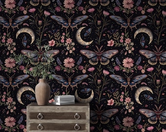 May include: A dark wallpaper pattern featuring repeating illustrations of moths, crescent moons, and pink flowers. In front of the wallpaper sits a wooden chest of drawers with two drawers, a ceramic vase with green foliage, and a stack of white books.