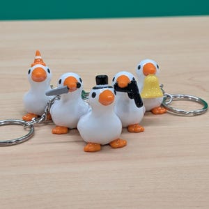 May include: A collection of five white goose figurines with orange beaks and feet, each holding a different accessory. One wears a top hat, another a party hat, and others hold a knife, a gun, and a bell. Each figurine is attached to a silver keychain ring.