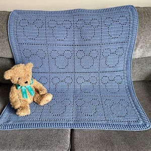 Op de afbeelding: Een blauwe gehaakte babydeken met een herhalend patroon van harten en sterren. De deken ligt op een grijze bank met een bruine teddybeer erop.