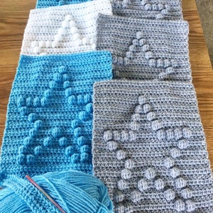 Op de afbeelding: Een close-up van gehaakte vierkanten in blauw en grijs met een bolletje blauwe garen en een haaknaald. De vierkanten hebben een reliëfpatroon in de vorm van een ster of boom.