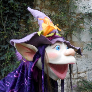 Può includere: Burattino viola e bianco con una grande bocca, capelli lunghi castani e un cappello viola con un fiore arancione. Il burattino ha grandi orecchie e occhi azzurri.