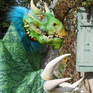 May include: A green dragon puppet with blue fur, white claws, and a toothy grin. The puppet is made of fabric and has a realistic appearance.