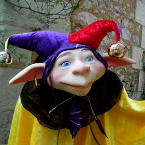 Puede incluir: Un gorro de bufón morado y rojo con campanas en los extremos de las puntas. El bufón lleva una capa amarilla y tiene una cara blanca con ojos azules.