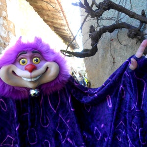 Peut inclure: Un costume en fourrure violet et noir avec un grand visage souriant et une cloche autour du cou. Le costume est porté par une personne avec une main levée en signe de salut.