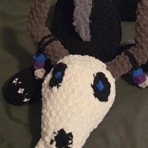 Peut inclure: Une peluche faite à la main en crochet en forme de crâne avec des cornes. Le crâne est blanc avec des détails noirs et bleus. Les cornes sont marron et la tête est noire. Le jouet est fait de fil doux et texturé.