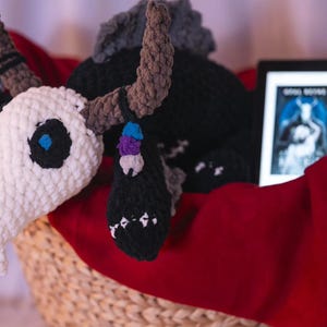 May include: A handmade crochet bull skull plushie with black, white, and brown details, resting in a woven basket lined with a red cloth. The skull has blue eyes and decorative accents. A digital tablet with a book cover is also in the basket.