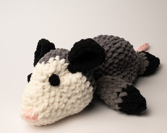 Könnte beinhalten: Ein gehäkeltes Plüschtier in Form eines Opossums. Das Opossum liegt auf der Seite mit dem Kopf auf dem Boden. Das Opossum ist weiß mit grauen und schwarzen Akzenten. Das Opossum hat eine rosa Nase und schwarze Augen.