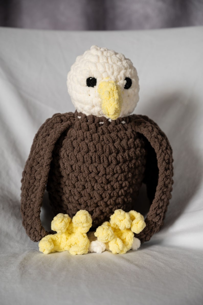 May include: A crocheted stuffed eagle toy with a white head, brown body, and yellow feet. The eagle has black eyes and a yellow beak.