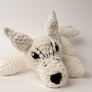 May include: White crocheted plush toy shaped like a wolf with black eyes and a black nose. The toy is lying on its side with its head turned to the side.