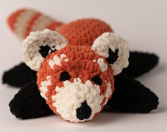 May include: A crocheted red panda plush toy with black eyes, a black nose, and a white and orange body. The toy is lying on its belly with its legs outstretched.