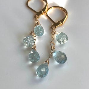 May include: Gold-tone leverback earrings with multiple light blue, faceted, teardrop-shaped gemstones dangling from delicate chains. The earrings are displayed against a white background.