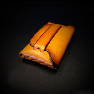 May include: A handcrafted, leather wallet in shades of tan and brown. The wallet features a flap closure and a decorative, textured edge. The leather has a natural, aged appearance, with darker edges and a lighter center.