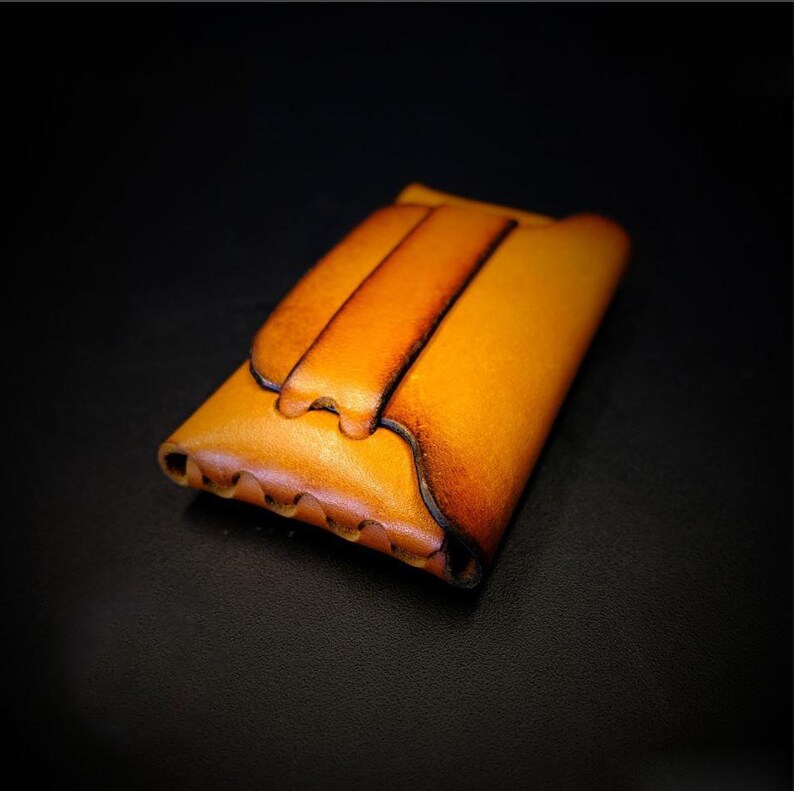 May include: A handcrafted, leather wallet in shades of tan and brown. The wallet features a flap closure and a decorative, textured edge. The leather has a natural, aged appearance, with darker edges and a lighter center.