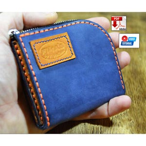 May include: A blue leather coin pouch with a zip closure, featuring orange stitching along the edges and a rectangular tan leather patch embossed with the text FIMAS. The design includes a PDF icon and a video tutorial icon in the corner.
