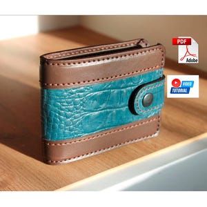 Puede incluir: Una cartera plegable con un panel central de textura de cocodrilo turquesa, enmarcado por cuero marrón. La cartera tiene un cierre de botón a presión y se muestra sobre una superficie de madera. El diseño incluye detalles cosidos.