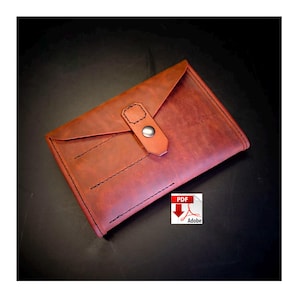 Puede incluir: Cartera de cuero marrón rojizo estilo sobre con cierre de solapa y broche metálico. La cartera presenta costuras visibles y un diseño minimalista.